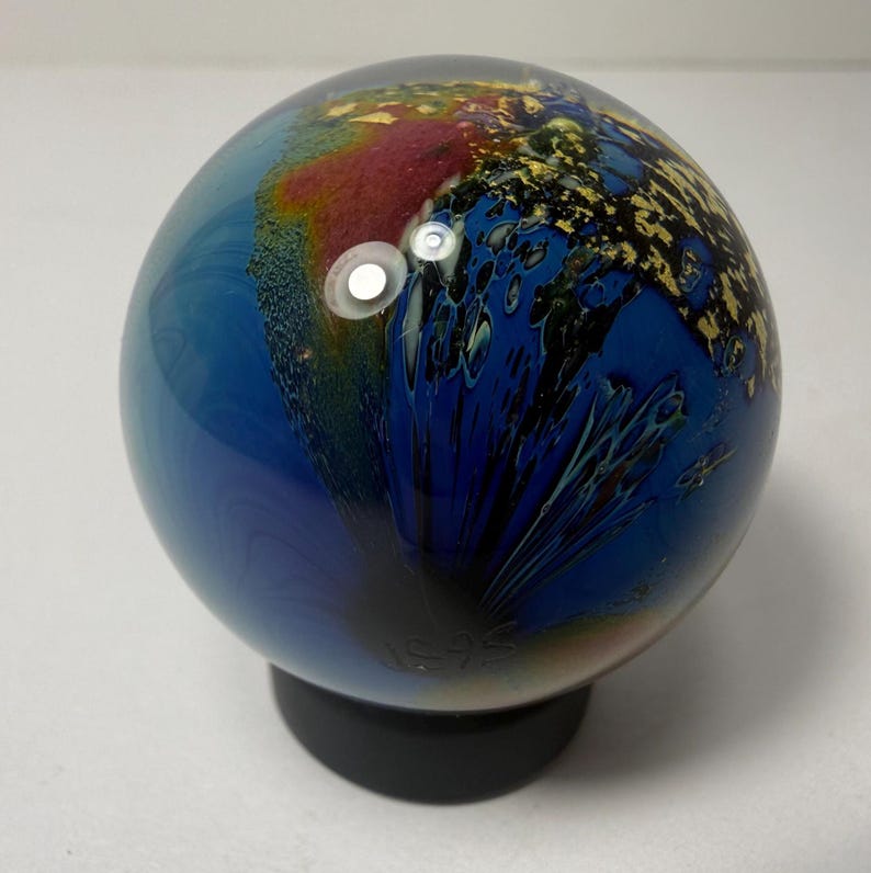 Puede incluir: Una esfera decorativa de vidrio con una vibrante mezcla de colores, incluyendo azul, rojo y dorado. La esfera se asienta sobre una base negra. El vidrio tiene un acabado brillante y un dise&ntilde;o abstracto &uacute;nico. Es una pieza de arte decorativa.