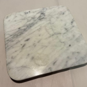May include: A square marble cutting board with rounded corners. The board has a white base with gray veining throughout. The surface is smooth and polished, suitable for food preparation or serving.