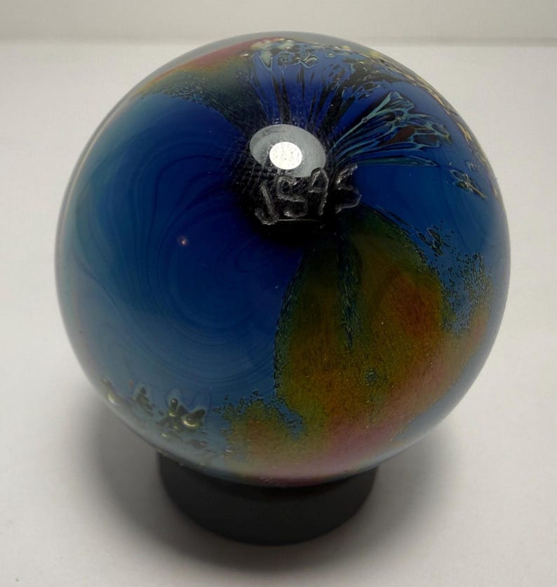 Puede incluir: Una esfera de vidrio decorativa con un patr&oacute;n arremolinado de tonos azules, naranjas y amarillos. La pieza de vidrio descansa sobre una base negra. El texto "JS45" est&aacute; grabado en el vidrio.