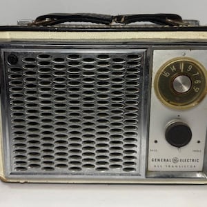 May include: Vintage General Electric All Transistor radio with a black leather handle. The radio has a silver speaker grill, a gold-toned dial, and a black control knob. The radio's casing is off-white with black trim.