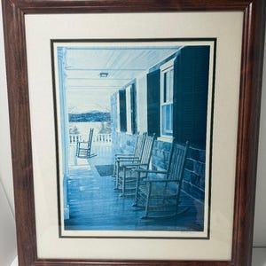 May include: A framed print depicting a porch with several wooden rocking chairs. The print has a blue and white color scheme, with a view of a lake and trees in the background. The frame is a dark brown color.