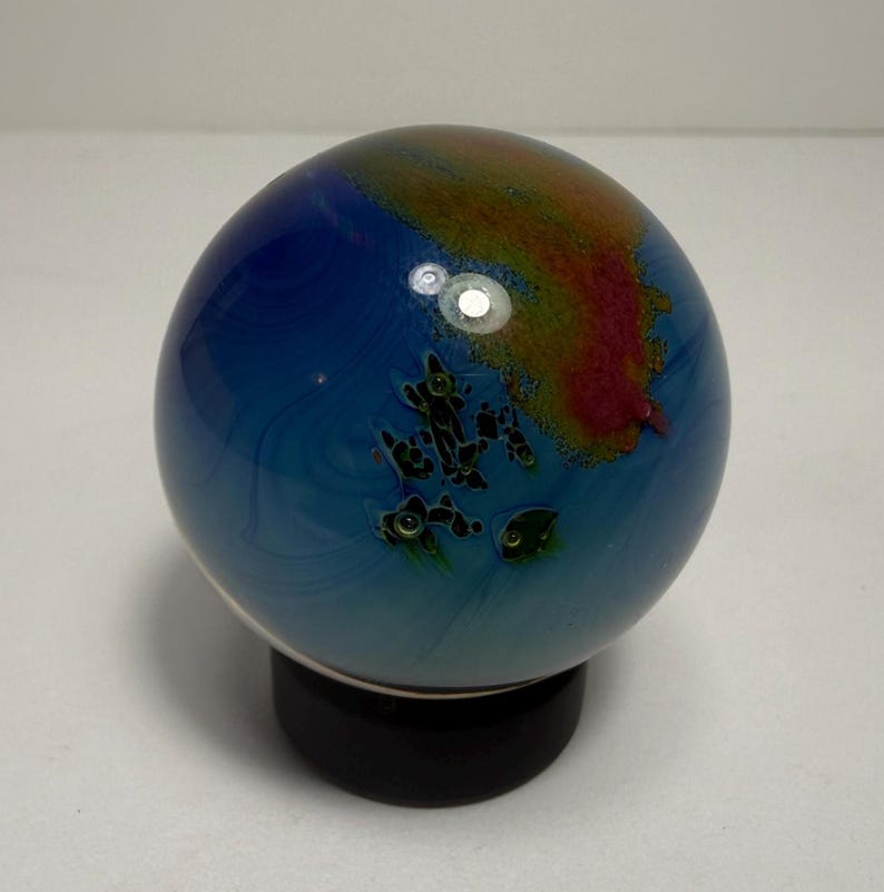 Puede incluir: Un globo de cristal decorativo con un patr&oacute;n arremolinado azul y verde, que se asemeja a un planeta. El globo tiene acentos rojos y marrones y se asienta sobre una base negra. La esfera de cristal es un art&iacute;culo decorativo &uacute;nico.