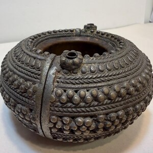 May include: An antique, round metal bowl with intricate, raised patterns. The bowl features a detailed design of small spheres, rope-like textures, and floral accents. A vertical metal band secures the bowl's side, and small decorative elements are present on the rim.