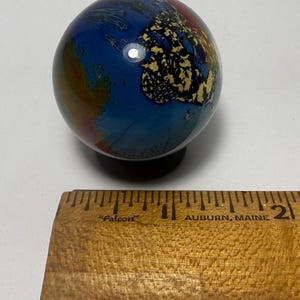 Puede incluir: Una esfera decorativa de vidrio con un patr&oacute;n arremolinado de colores azul, dorado, rojo y marr&oacute;n. La esfera tiene aproximadamente 5 cm de di&aacute;metro y se asienta sobre una peque&ntilde;a base negra. La esfera est&aacute; junto a una regla de madera.