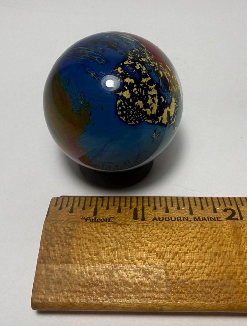 Puede incluir: Una esfera decorativa de vidrio con un patr&oacute;n arremolinado de colores azul, dorado, rojo y marr&oacute;n. La esfera tiene aproximadamente 5 cm de di&aacute;metro y se asienta sobre una peque&ntilde;a base negra. La esfera est&aacute; junto a una regla de madera.