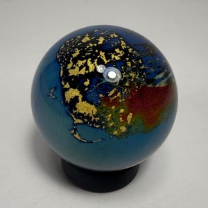 Puede incluir: Un globo terr&aacute;queo decorativo de vidrio con un dise&ntilde;o de mapa. Presenta oc&eacute;anos azules, continentes dorados y detalles en rojo y naranja. El globo est&aacute; montado sobre una base negra, ofreciendo una representaci&oacute;n art&iacute;stica &uacute;nica del mundo.