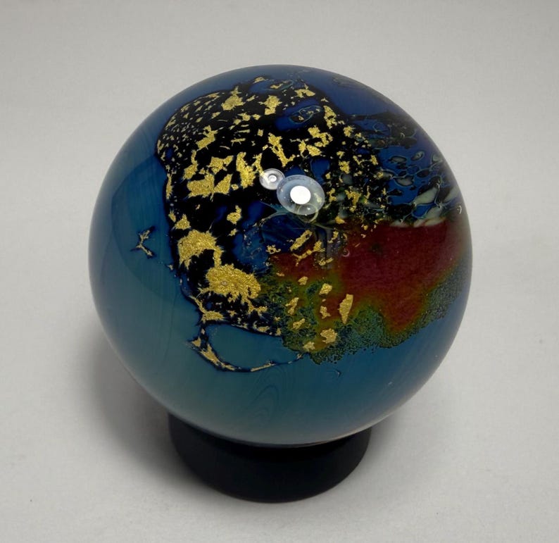 Puede incluir: Un globo terr&aacute;queo decorativo de vidrio con un dise&ntilde;o de mapa. Presenta oc&eacute;anos azules, continentes dorados y detalles en rojo y naranja. El globo est&aacute; montado sobre una base negra, ofreciendo una representaci&oacute;n art&iacute;stica &uacute;nica del mundo.