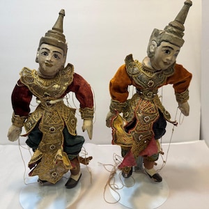 May include: Two antique marionette puppets. Each puppet has a painted face, a conical headdress, and is dressed in ornate, gold-trimmed costumes with red and orange velvet sleeves. The puppets are suspended by strings and stand on clear plastic bases.