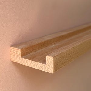 May include: A natural wood floating shelf with a grooved design. The shelf is mounted on a light peach-colored wall. The shelf is approximately 3 inches wide and 1 inch thick.