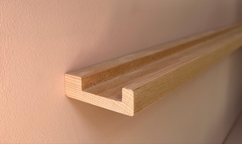May include: A natural wood floating shelf with a grooved design. The shelf is mounted on a light peach-colored wall. The shelf is approximately 3 inches wide and 1 inch thick.