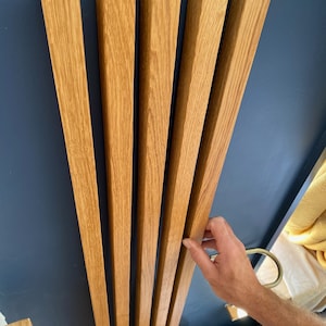 Peut inclure: Ensemble de cinq lattes en bois clair, probablement pour un dossier de chaise ou un élément mural décoratif, contre un mur bleu foncé. Le bois clair présente un grain visible. Le design est moderne et minimaliste.