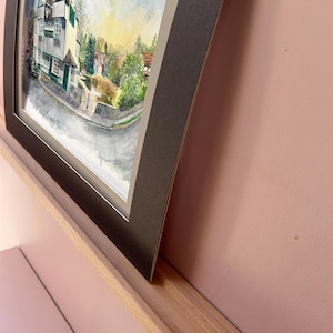 May include: Framed watercolor painting of a village scene. The artwork depicts a white and green building with a red roof, trees, and a road. The painting is set in a black frame with a white mat, displayed on a wooden shelf against a pink wall.
