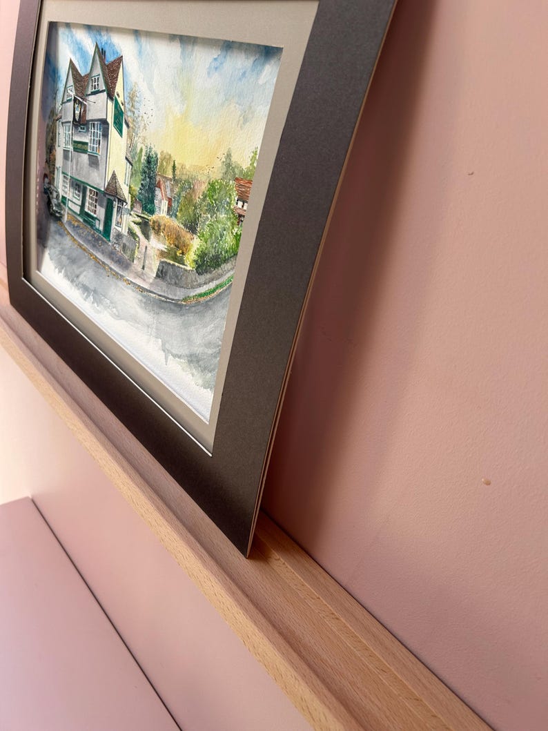 May include: Framed watercolor painting of a village scene. The artwork depicts a white and green building with a red roof, trees, and a road. The painting is set in a black frame with a white mat, displayed on a wooden shelf against a pink wall.