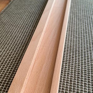 May include: A long, unfinished wooden tray with a recessed channel. The tray is made of light-colored wood and has a rectangular shape. The tray is designed to hold items and is placed on a textured surface.