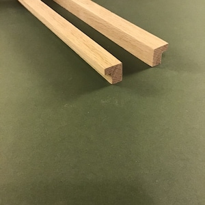 May include: Two pieces of wooden trim with a 45-degree angle cut on one end. The trim is light brown in colour and is on a green surface.