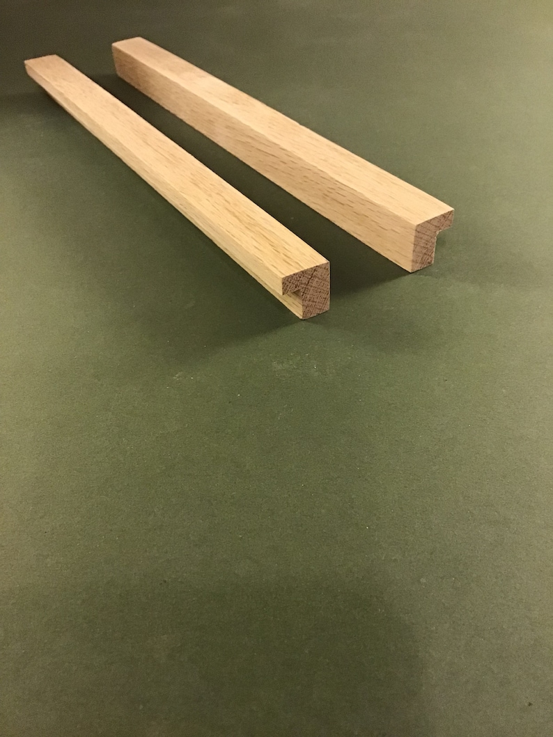 May include: Two pieces of wooden trim with a 45-degree angle cut on one end. The trim is light brown in colour and is on a green surface.