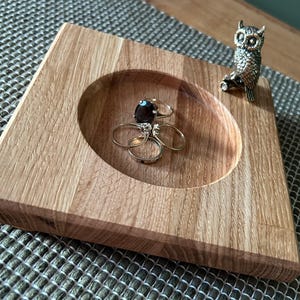 May include: A wooden ring dish with a circular recess, containing rings with gemstones. A small owl ornament is positioned on the edge. The dish is made of light-coloured wood with a visible grain pattern.