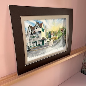 May include: Watercolor painting of a building with a sign that reads "The King's Garden". The building is white and green with a brown roof. The painting is framed in a dark brown mat and frame.