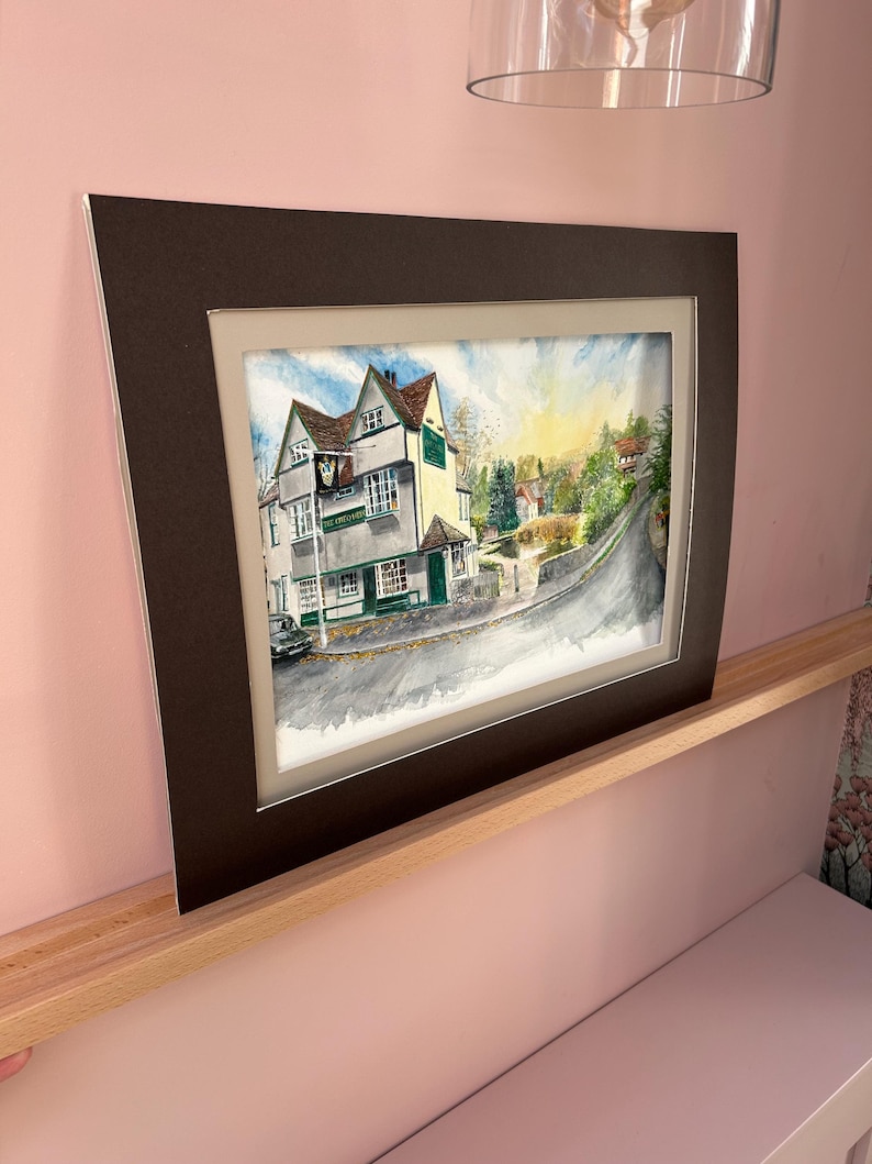 May include: Watercolor painting of a building with a sign that reads "The King's Garden". The building is white and green with a brown roof. The painting is framed in a dark brown mat and frame.