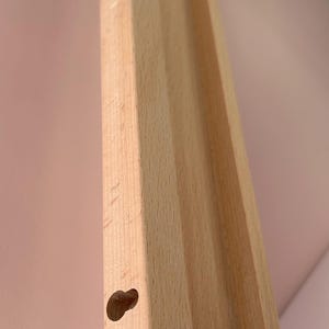 May include: Close-up of a light-colored wooden furniture leg with a rounded hole. The leg has a grooved design and is angled towards the top right. The background is a soft pink.