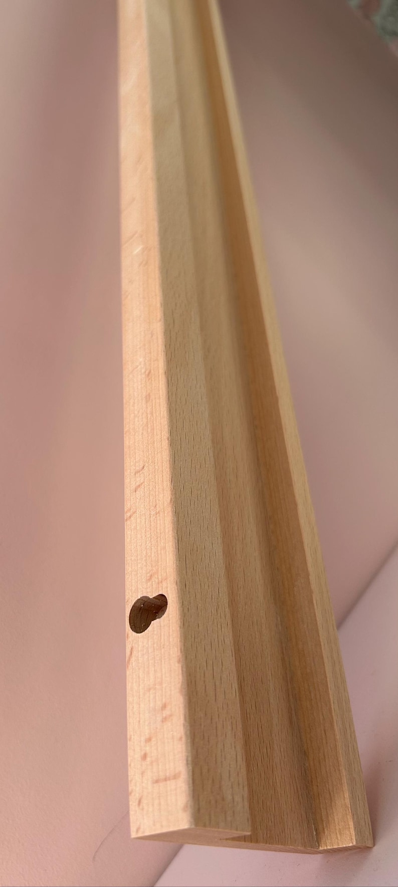 May include: Close-up of a light-colored wooden furniture leg with a rounded hole. The leg has a grooved design and is angled towards the top right. The background is a soft pink.