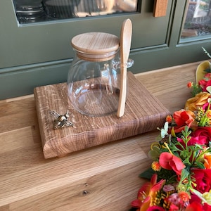 May include: A clear glass jar with a wooden lid and spoon sits on a wooden board. A brass bee decoration is attached to the board. The jar is empty. The background features a green door and a floral display. The jar is for serving.