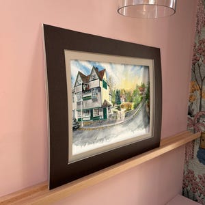 May include: Watercolor painting of a building with a green door and sign, set on a street with a curved road. The artwork is framed in a dark brown mat and frame. The painting depicts a sunny day with trees and other buildings in the background.