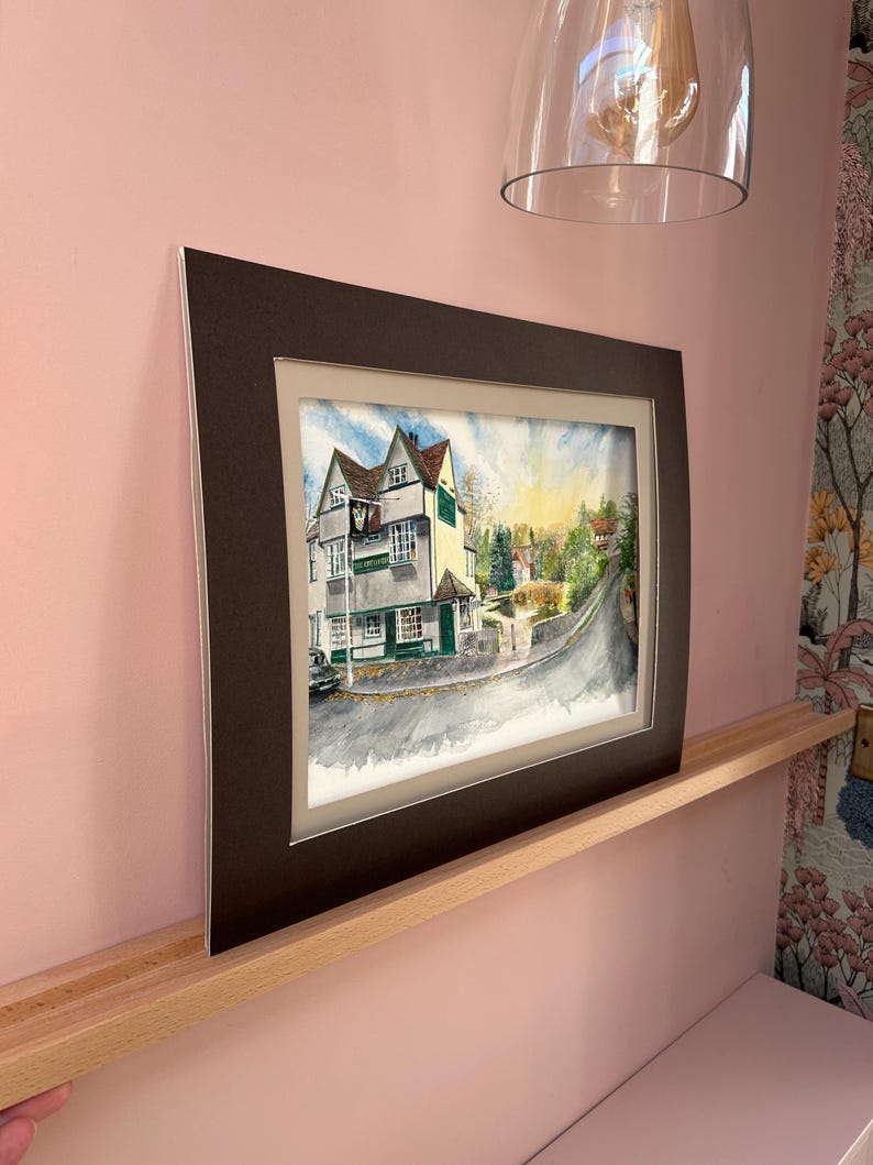May include: Watercolor painting of a building with a green door and sign, set on a street with a curved road. The artwork is framed in a dark brown mat and frame. The painting depicts a sunny day with trees and other buildings in the background.