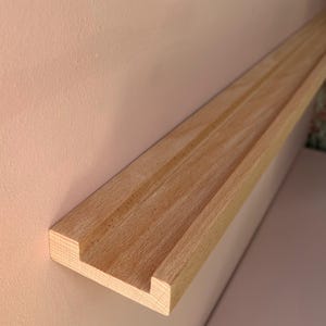 May include: A light-colored wooden shelf with a grooved design, mounted on a pale pink wall. The shelf has a rectangular profile with a recessed channel running along its length. The wood grain is visible.