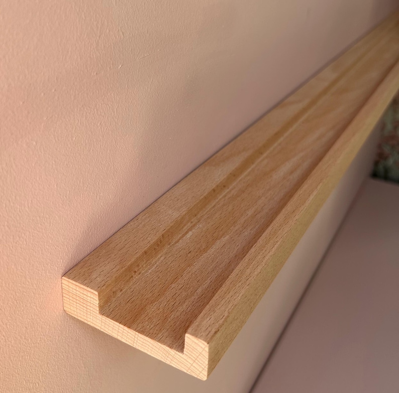 May include: A light-colored wooden shelf with a grooved design, mounted on a pale pink wall. The shelf has a rectangular profile with a recessed channel running along its length. The wood grain is visible.