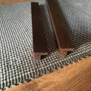 Peut inclure: Deux poignées de meubles en bois brun foncé, de forme rectangulaire avec un petit rebord à une extrémité. Elles reposent sur un dessous de plat texturé gris et blanc sur une surface en bois. Chaque poignée mesure environ 20 cm de long.