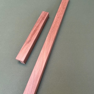 Peut inclure: Deux pièces de bois rectangulaires, une longue et une courte, de couleur brun rougeâtre, sont disposées en diagonale sur un fond vert sourd. Le grain du bois est visible, ajoutant de la texture aux formes géométriques simples.