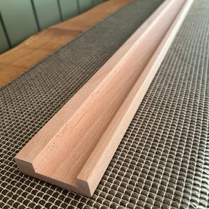 May include: A long, unfinished wooden tray with a recessed channel. The tray is made of light-colored wood and has a rectangular shape. The tray is designed to hold items and is placed on a textured surface.
