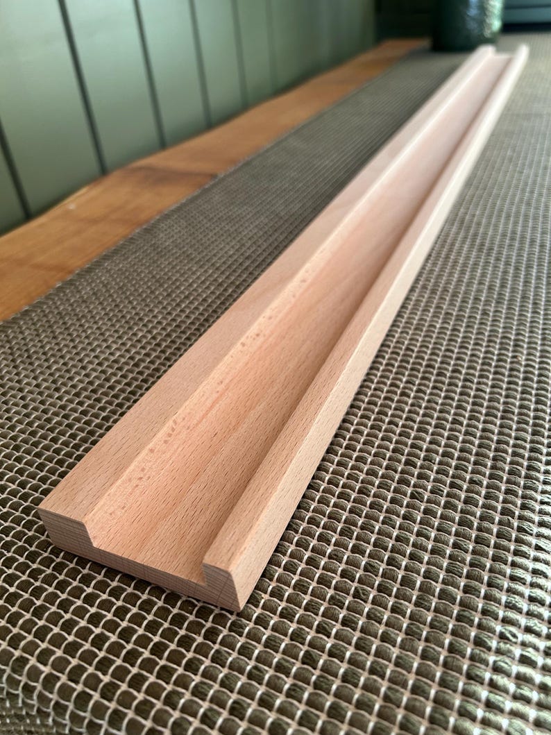 May include: A long, unfinished wooden tray with a recessed channel. The tray is made of light-colored wood and has a rectangular shape. The tray is designed to hold items and is placed on a textured surface.