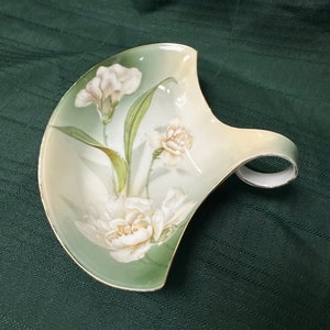 May include: A white porcelain dish with a green and white floral design. The dish has a handle and is shaped like a crescent moon.