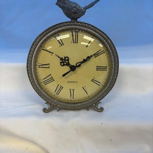 May include: A round quartz table clock with a metallic bronze-toned frame featuring a decorative bird figurine on top. The clock face is cream-colored with black Roman numerals and ornate black hands. The base has two small, curved metal feet.