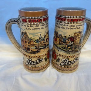 May include: Two ceramic beer steins featuring a tan base with a relief illustration of a German village, a bridge, and a tower. The text reads THE STROH BREWERY COMPANY and Stroh's. Each stein has a curved handle and a metallic-toned rim at the top.
