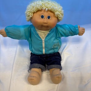 Puede incluir: Una muñeca Cabbage Patch Kids con cabello de lana rubio rizado y ojos azules. La muñeca viste una chaqueta cortavientos azul brillante con cremallera y el logotipo de Cabbage Patch Kids en el pecho, una camiseta a rayas amarillas y blancas y pantalones cortos de mezclilla azul oscuro.
