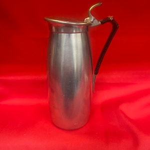 Puede incluir: Una jarra de plata con asa negra y tapa. La jarra tiene una superficie lisa y brillante.