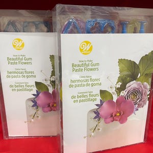 Może przedstawiać: Dwa zestawy kwiatów z gum paste Wilton. Każdy przezroczysty pakiet ma białą etykietę z logo Wilton i tekstem "How to Make Beautiful Gum Paste Flowers" w języku angielskim, hiszpańskim i francuskim. Ozdobione kwiatami z gum paste.