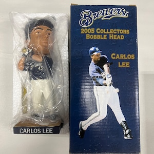 May include: A bobblehead figurine of baseball player Carlos Lee in a Brewers uniform, packaged in a box. The figurine is in a batting pose, with the name "CARLOS LEE" on the base. The box reads "2005 COLLECTORS BOBBLE HEAD".