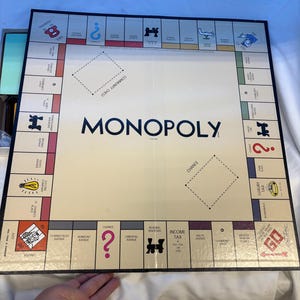 May include: A square Monopoly board game featuring a cream-colored center with the word Monopoly printed in black. The perimeter contains colored property spaces, railroad icons, Chance and Community Chest squares, and corner spaces like Go.