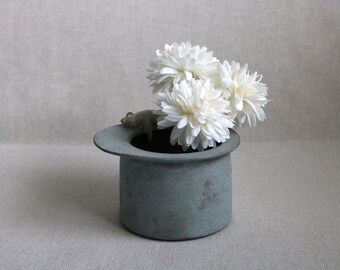 May include: A small, cylindrical, blue-green ceramic vase with a wide brim, holding three white, fluffy flowers. A small, grey frog figurine sits on the brim of the vase. The vase has a rustic, textured appearance.