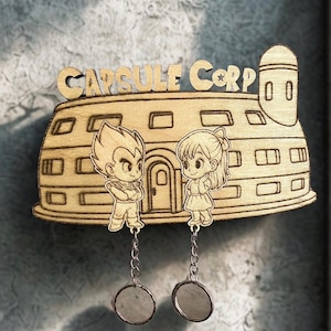 May include: A light-colored wooden wall decoration shaped like the Capsule Corp building, featuring engraved cartoon characters of a spiky-haired figure and a girl with a ponytail. Two small circular mirrors hang from the base by silver chains.