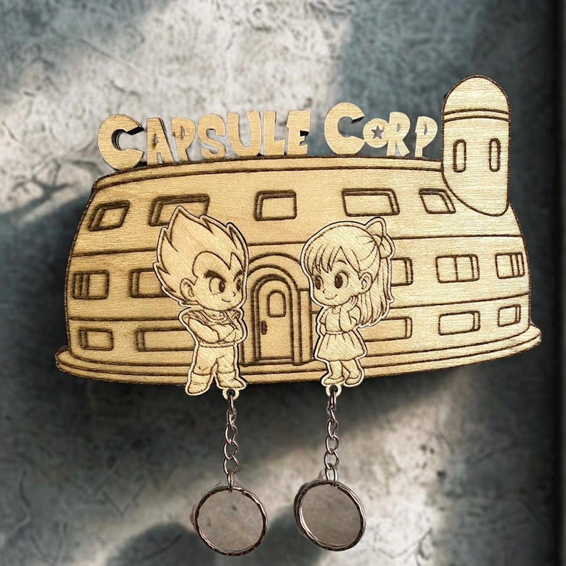 May include: A light-colored wooden wall decoration shaped like the Capsule Corp building, featuring engraved cartoon characters of a spiky-haired figure and a girl with a ponytail. Two small circular mirrors hang from the base by silver chains.