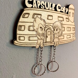 May include: A light-colored wooden wall-mounted key holder featuring a laser-engraved Capsule Corp building with two cartoon characters standing in front. Two silver metal chains with circular key rings hang from the bottom of the wooden piece.