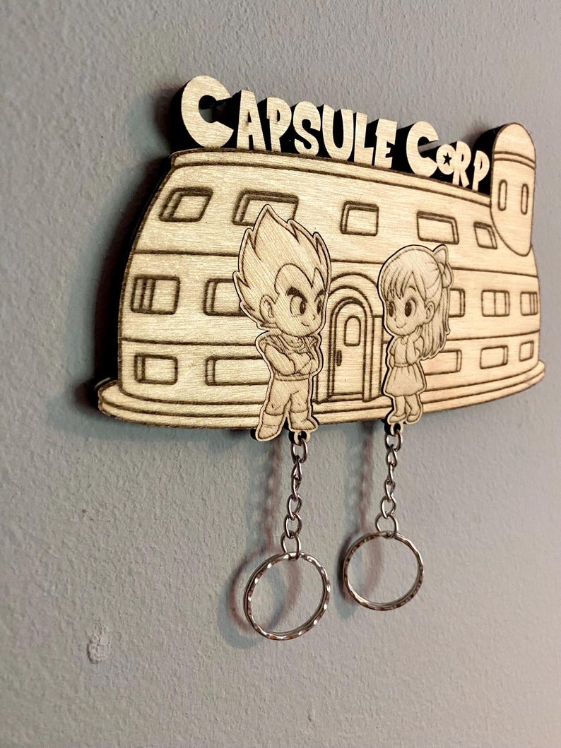May include: A light-colored wooden wall-mounted key holder featuring a laser-engraved Capsule Corp building with two cartoon characters standing in front. Two silver metal chains with circular key rings hang from the bottom of the wooden piece.