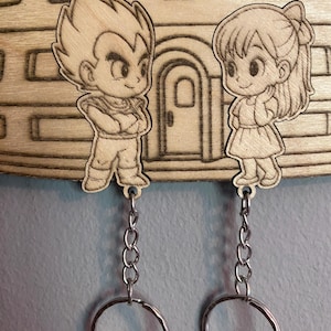 May include: Two wooden keychains featuring laser-engraved anime characters, a male figure with spiky hair and a female figure with a ponytail, both attached to silver metal chains and circular rings, mounted on a wooden base with a building design.