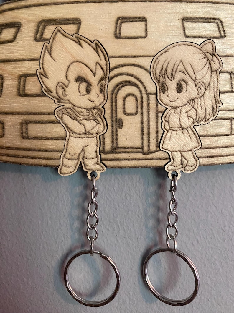 May include: Two wooden keychains featuring laser-engraved anime characters, a male figure with spiky hair and a female figure with a ponytail, both attached to silver metal chains and circular rings, mounted on a wooden base with a building design.