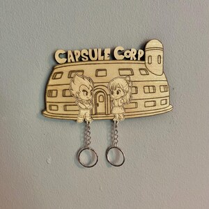 May include: A light-colored wooden wall-mounted key holder shaped like the Capsule Corp building. It features engraved anime-style characters and the text "CAPSULE CORP" at the top. Two silver metal chains with key rings hang from the bottom.